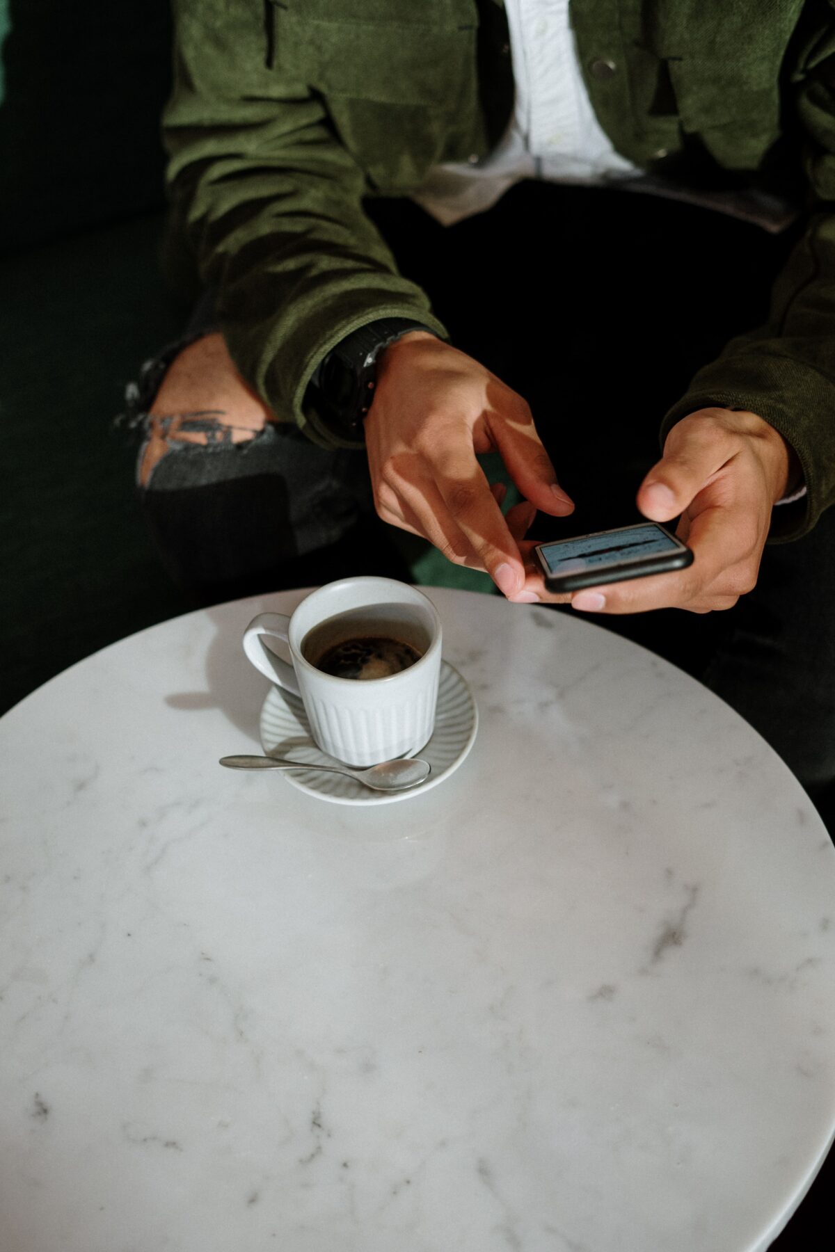 A person in a green jacket uses a smartphone while sitting at a white marble table with a cup of coffee and a spoon on a saucer. Only the person’s hands and part of their outfit are visible.